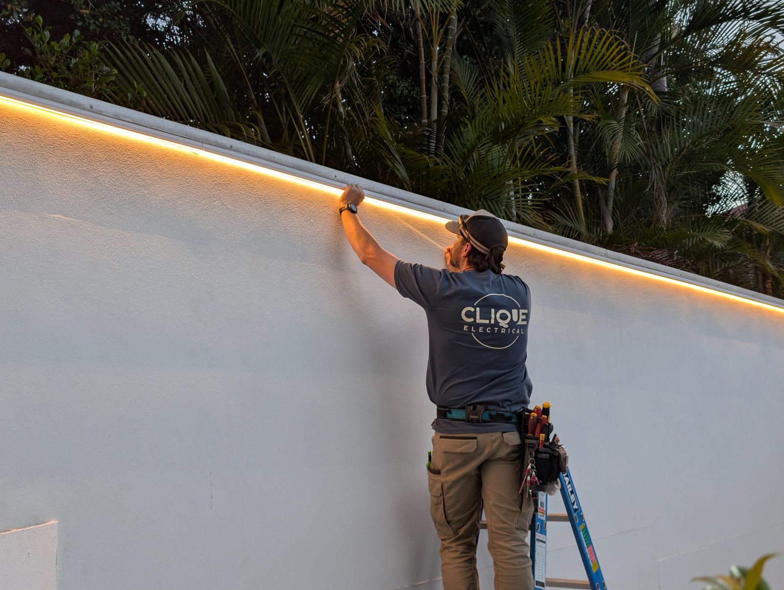 Clique Electrical electrician fitting warm white LED strip lighting along a rendered feature wall at dusk, South East Queensland